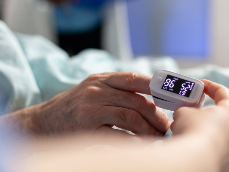 Close up of oxymeter attached on sick senior patient laying in hospital bed, monitoring hemoglobin. Medical staff using modern technology for reading blood oxygen saturation.